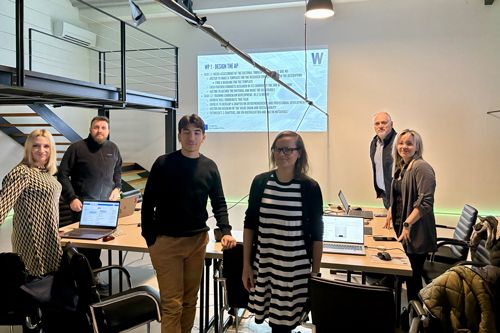 6 people standing around a U shaped table cluster, looking at the camera, posing. There is apresentation on the wall behind them