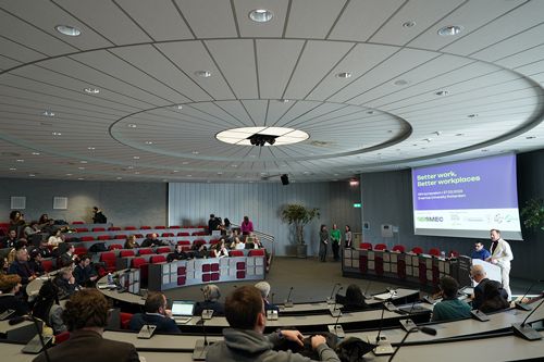 Round conference table with red chairs, people sitting at the tables. There is a person giving a presentation